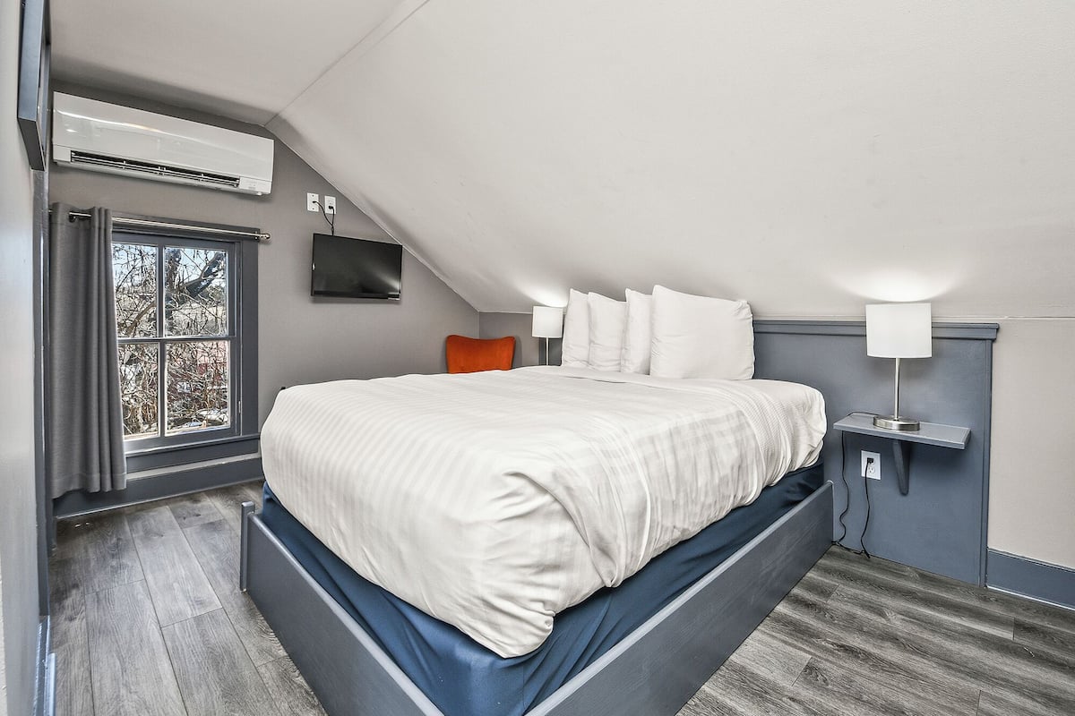 A modern bedroom with a sloped ceiling, a large bed with white bedding, a TV mounted on the wall, and a window with a view of trees.