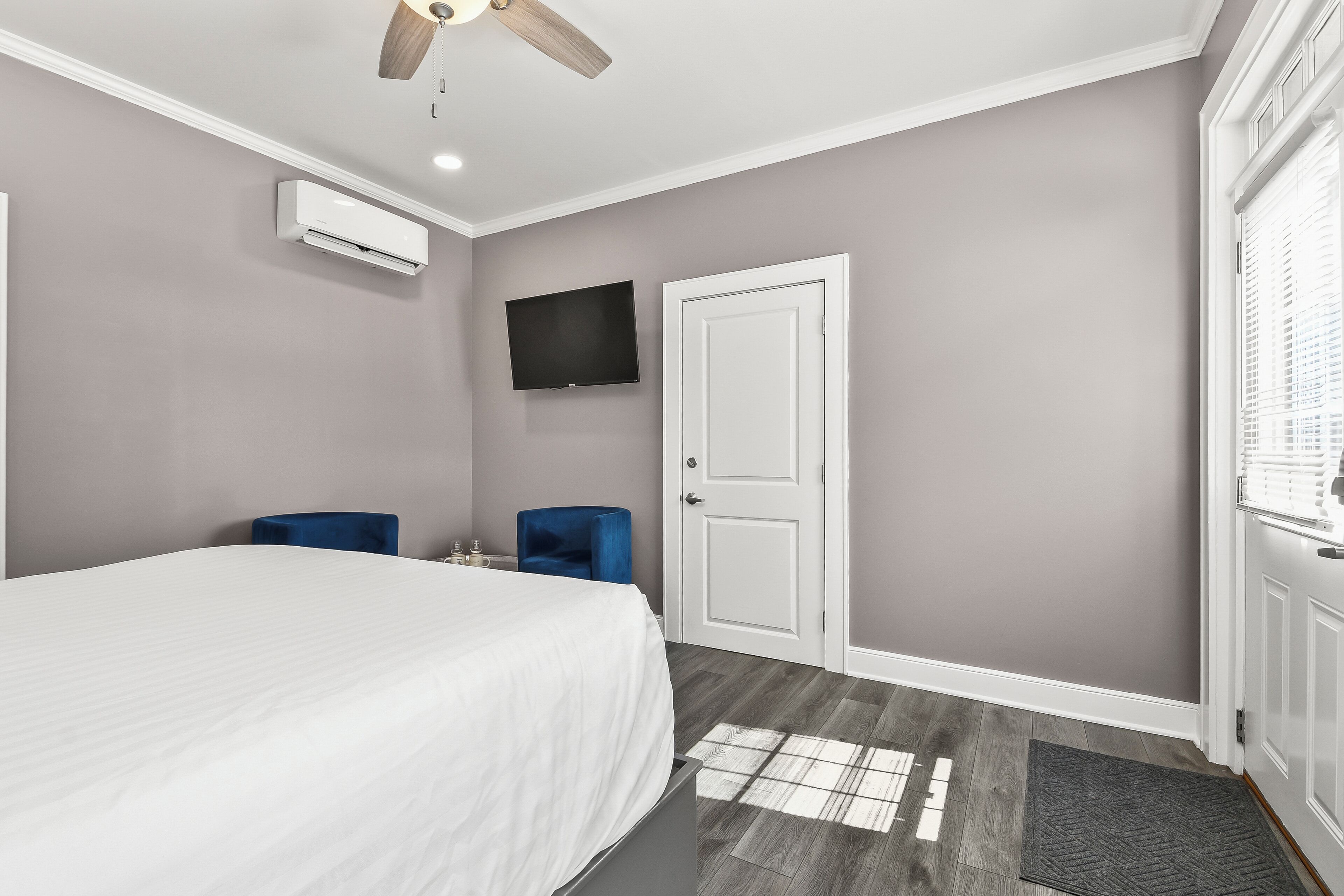 A bedroom with a bed, a ceiling fan, a TV, a door, and a window with blinds.