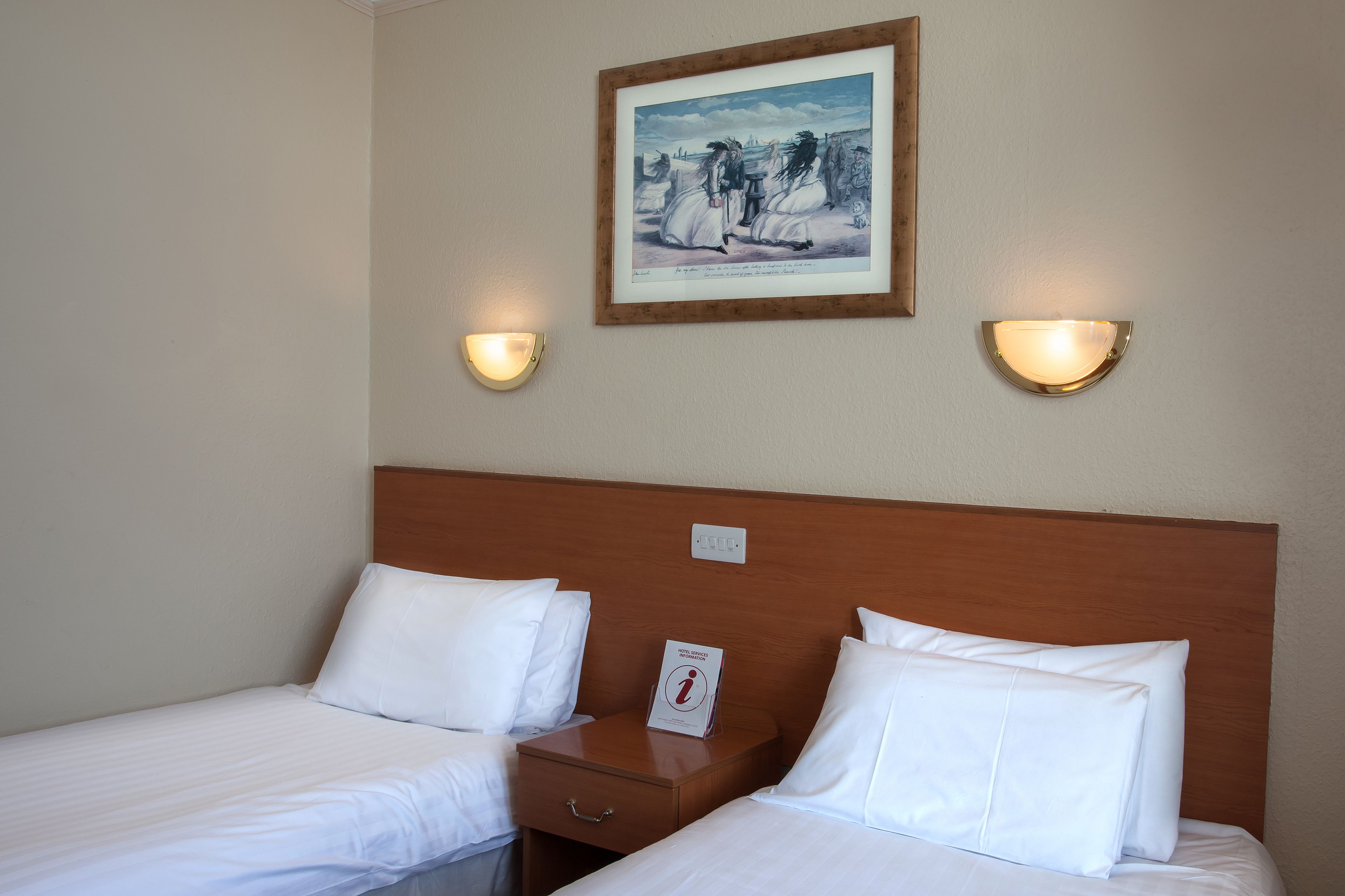 Habitación de hotel con dos camas, un cabecero de madera, una mesita de noche con un libro y apliques de pared.