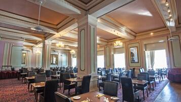 A spacious banquet hall with high ceilings, ornate moldings, and chandeliers. Tables are set with fine china and silverware.