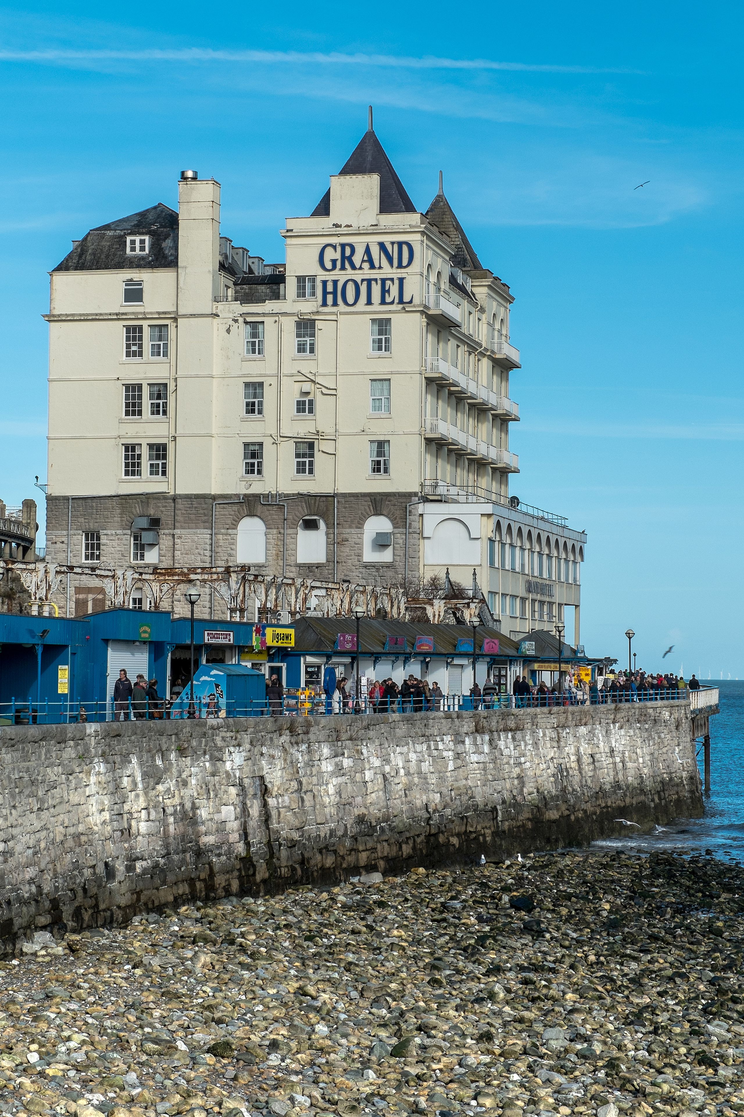 A large hotel with multiple floors and balconies, located near a waterfront with a rocky shore.