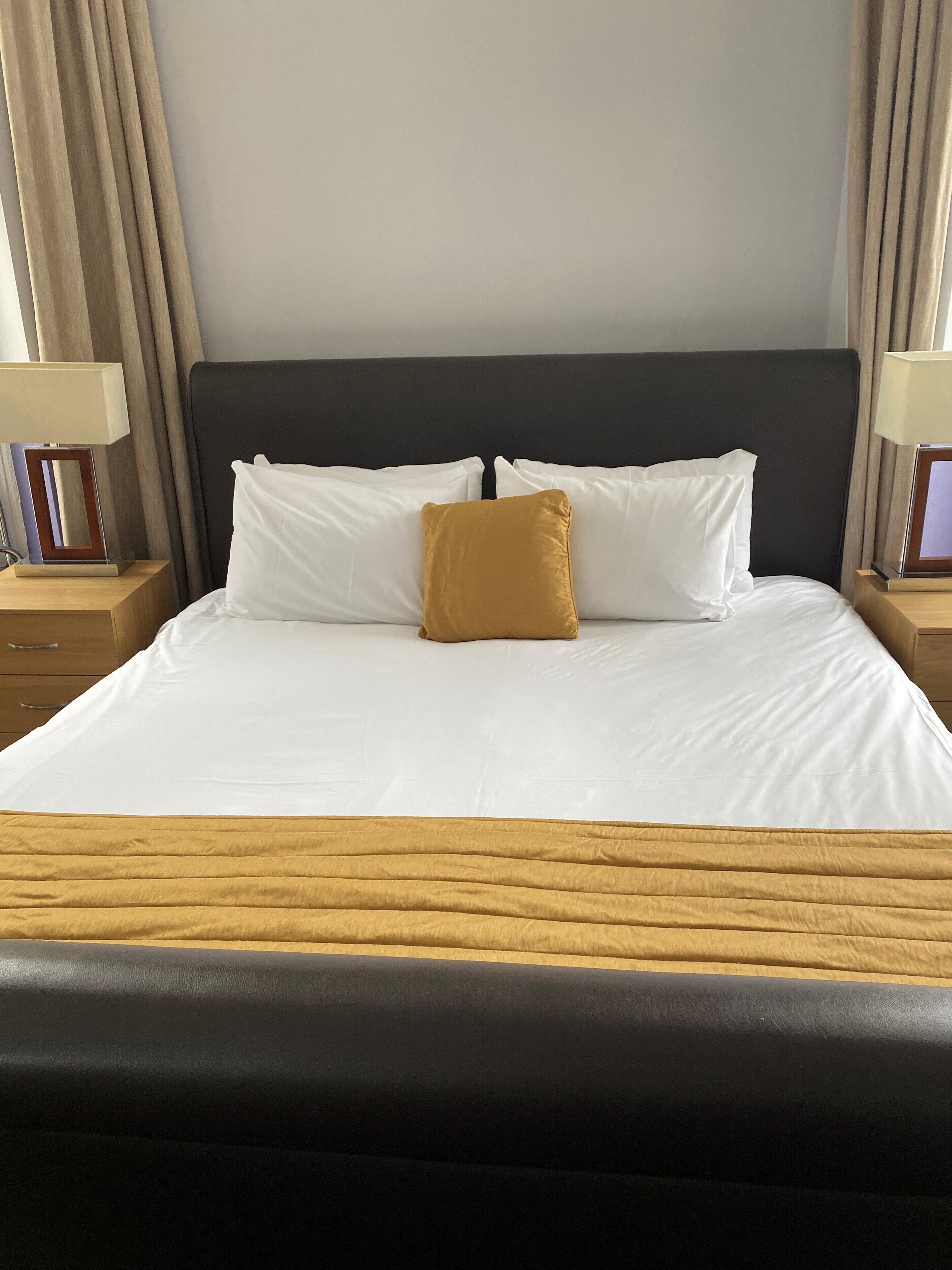 A neatly made bed with white and mustard pillows, a wooden headboard, and a wooden bedside table with a lamp.