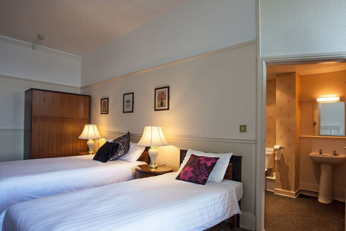 Une chambre d’hôtel avec deux lits, des lampes de chevet, une armoire, une salle de bain avec un lavabo et un miroir, et des tableaux encadrés accrochés au mur.