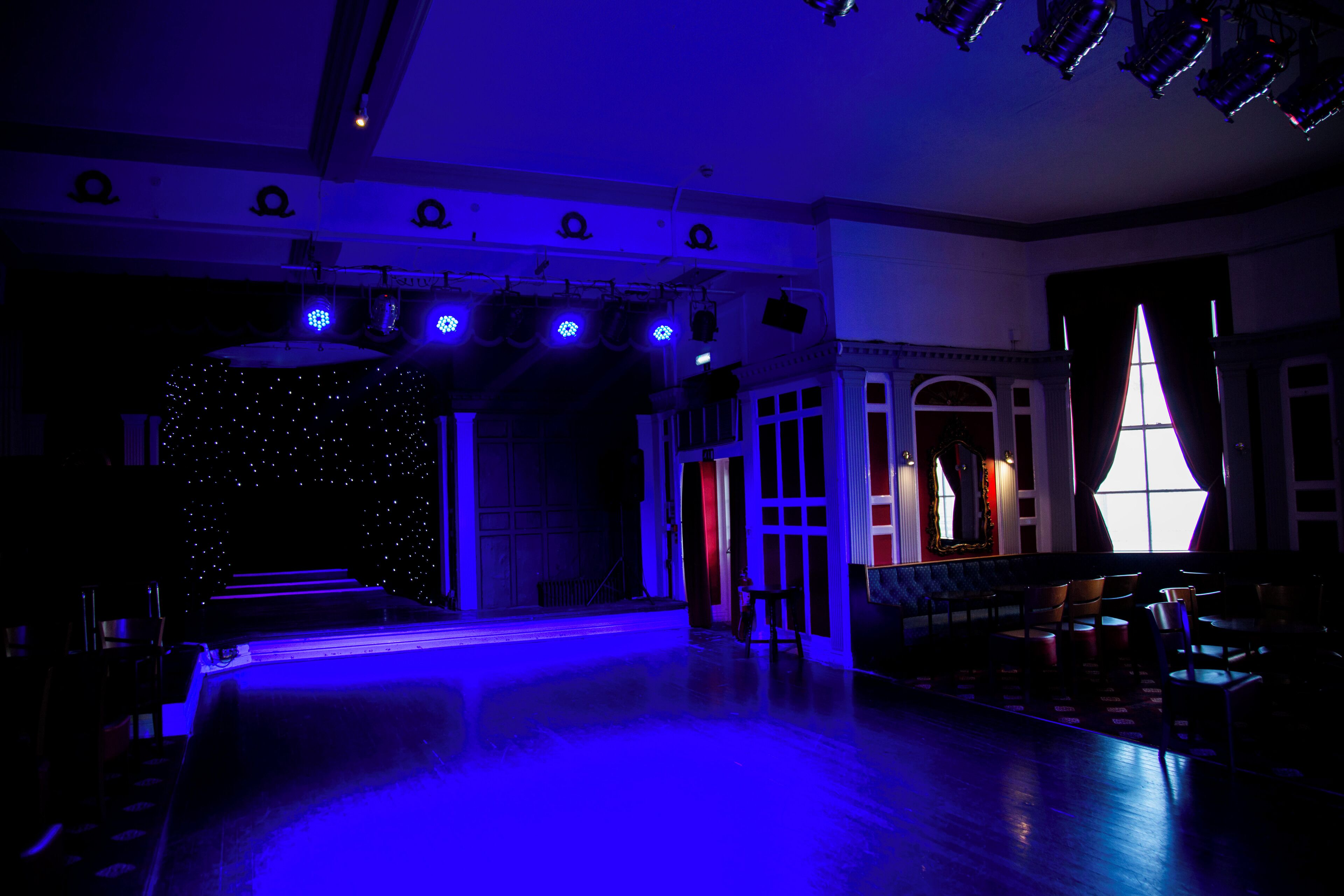 Una sala tenuemente iluminada con un escenario, zona de asientos y un ventanal con cortinas.