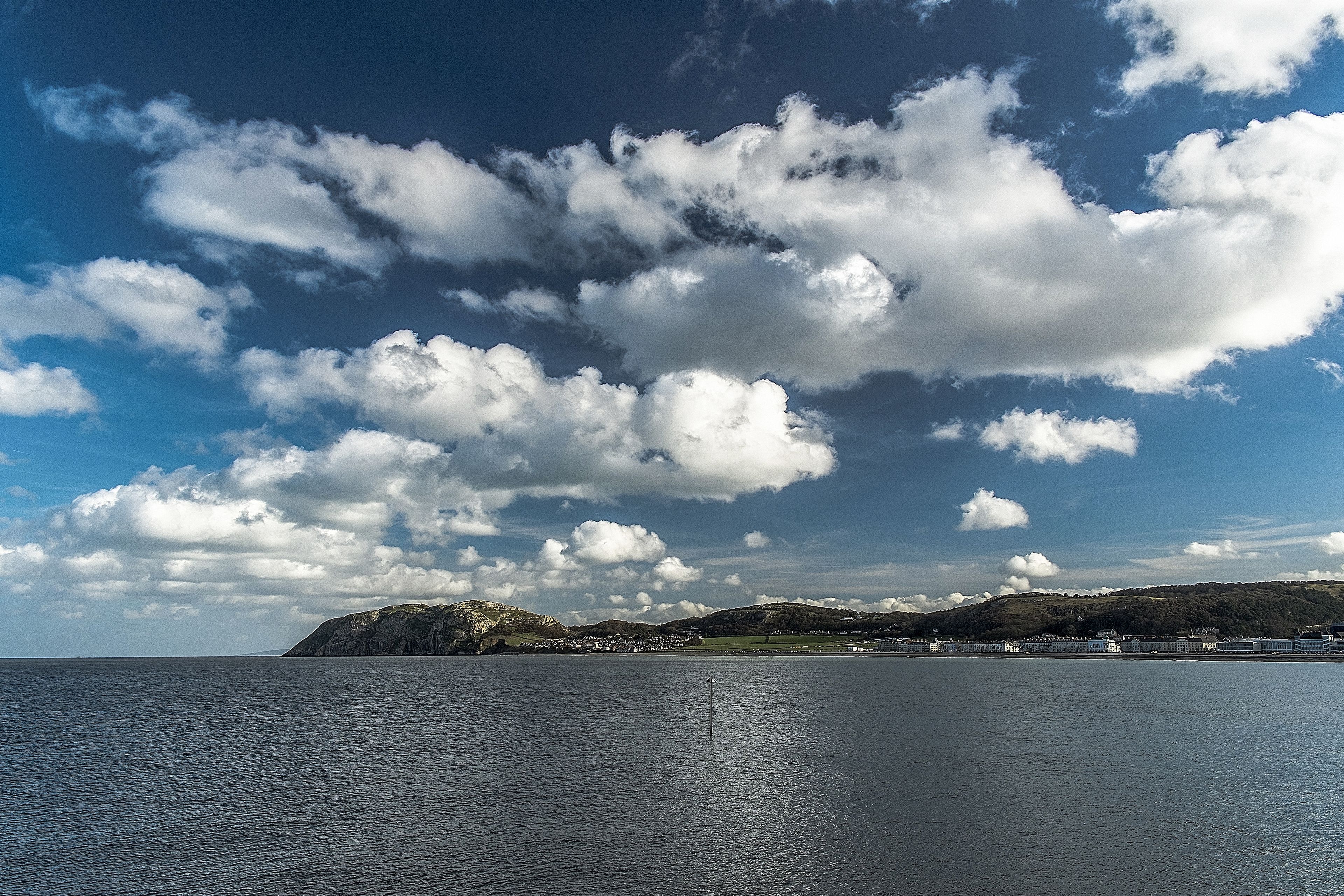 A coastal landscape with a calm sea, a clear sky with scattered clouds, and a hilly terrain in the background.