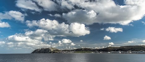A coastal landscape with a calm sea, a clear sky with scattered clouds, and a hilly terrain in the background.