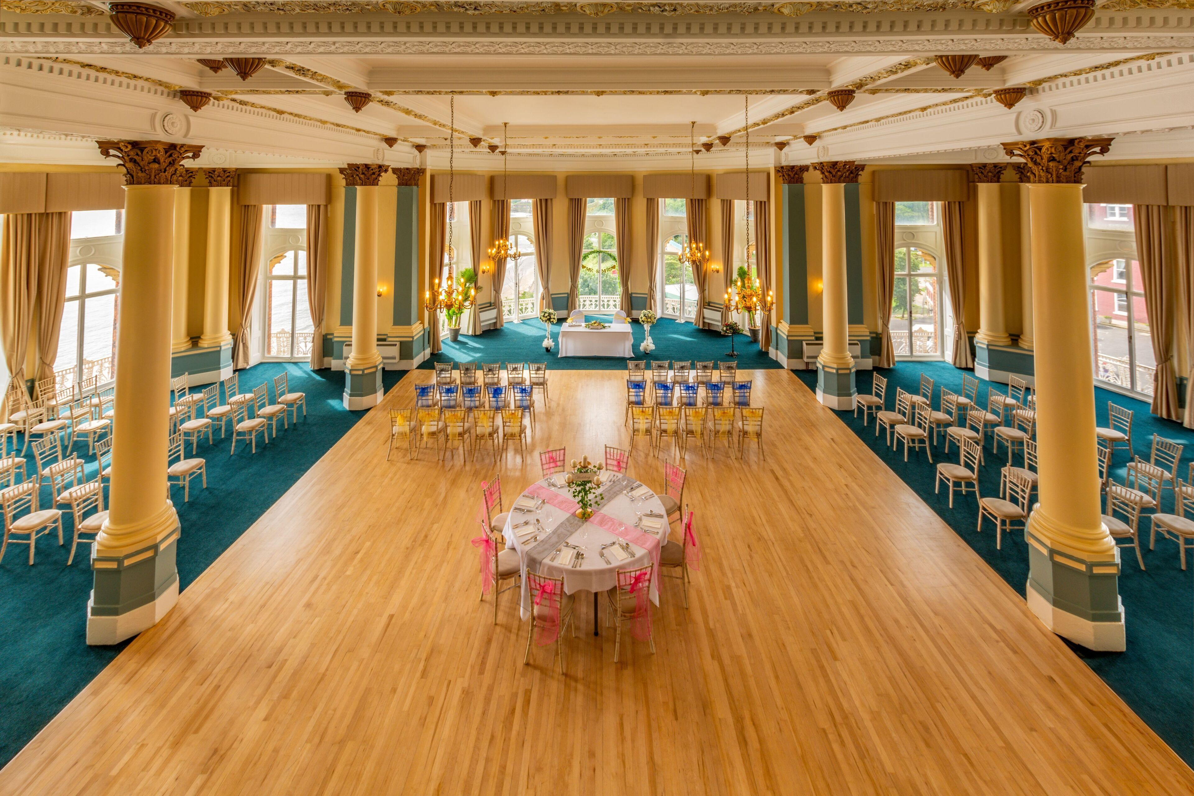 A large banquet hall with a long table set for a formal event, surrounded by chairs and columns.