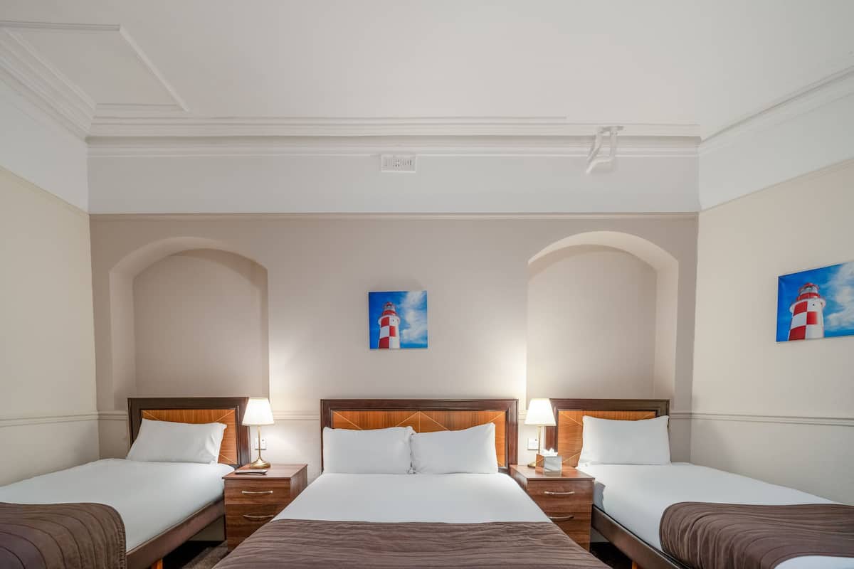 Une chambre d’hôtel avec trois lits, une tête de lit en bois, un tableau représentant un phare et un plafond blanc.