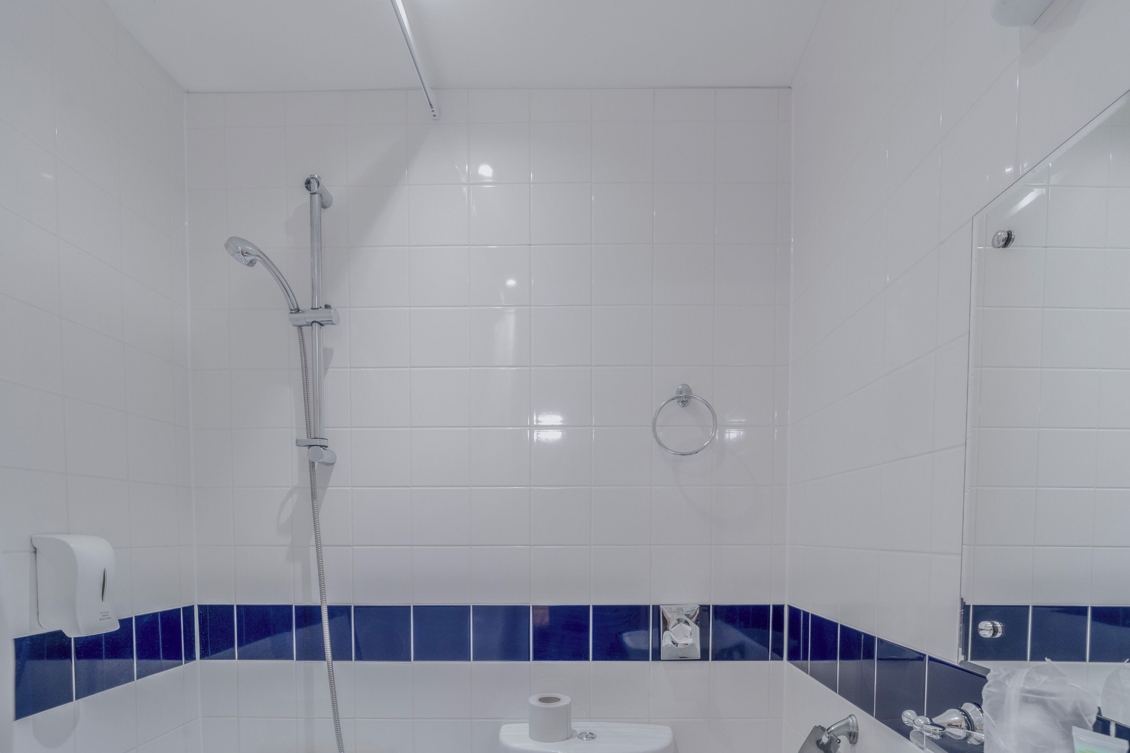 A modern bathroom with white tiles, a blue accent border, a shower with a handheld showerhead, a toilet, and a sink with a mirror.