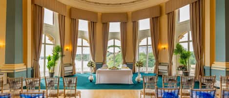 Salón de ceremonia de boda con una mesa central, sillas adornadas con lazos azules, ventanas amplias y vistas a zonas verdes.