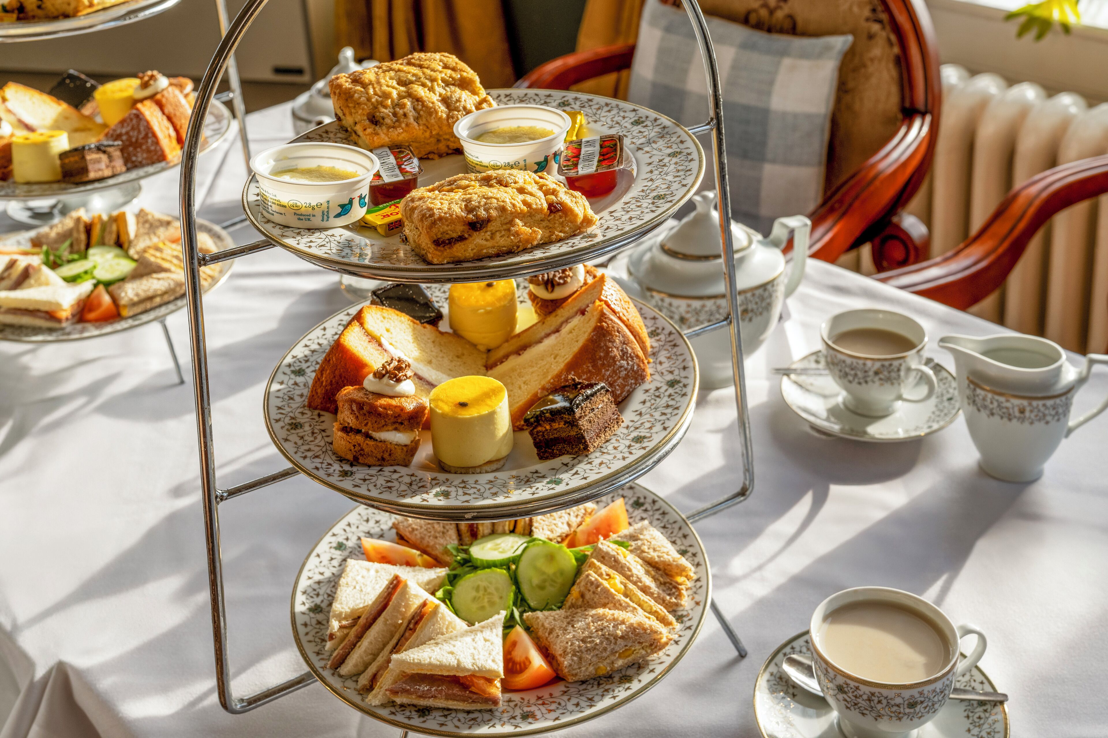 A tiered tray with sandwiches, scones, and butter, alongside cups of tea and a teapot on a table.
