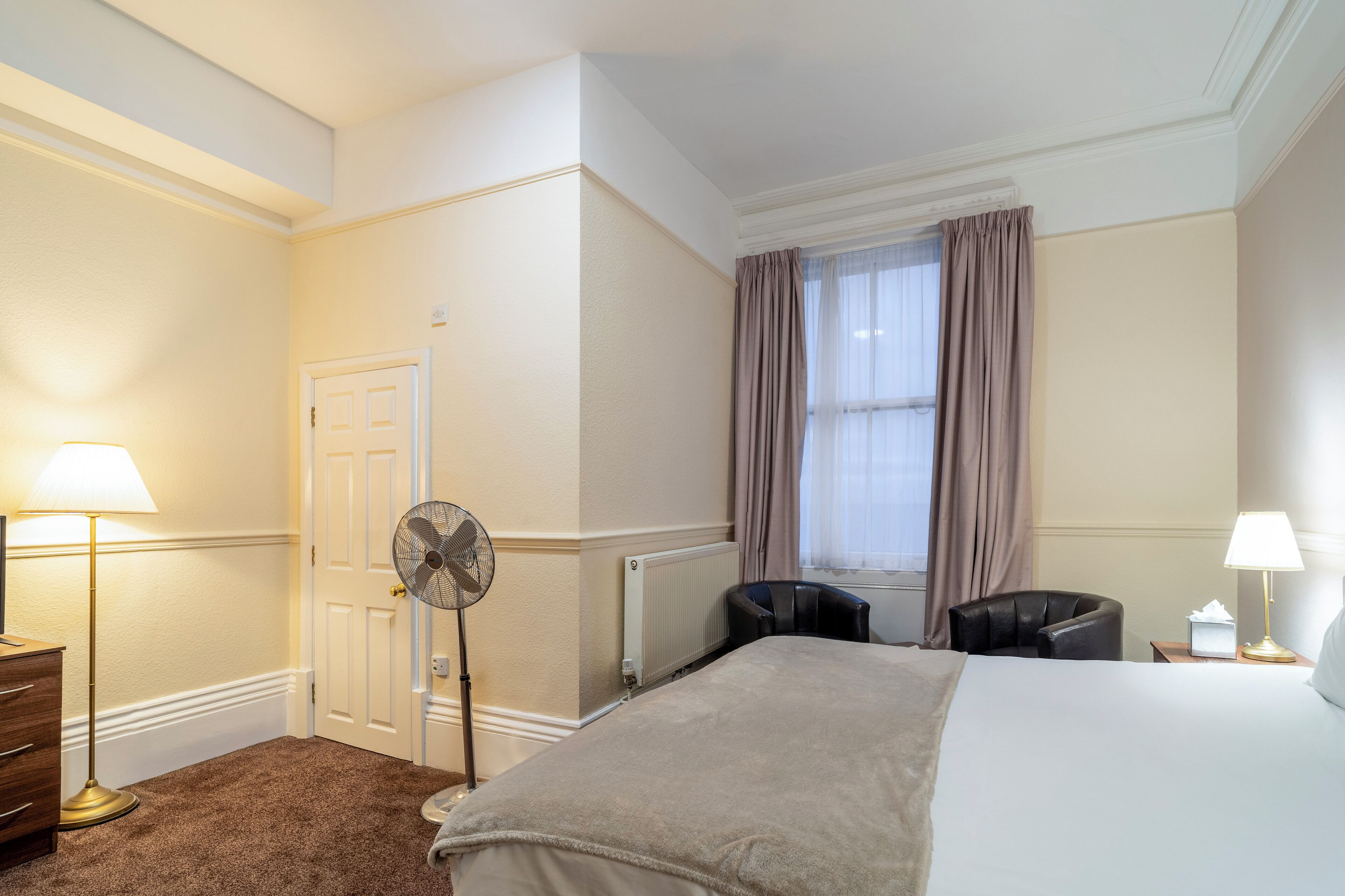 A hotel room with a bed, a desk, a chair, a fan, a lamp, and a window with curtains.