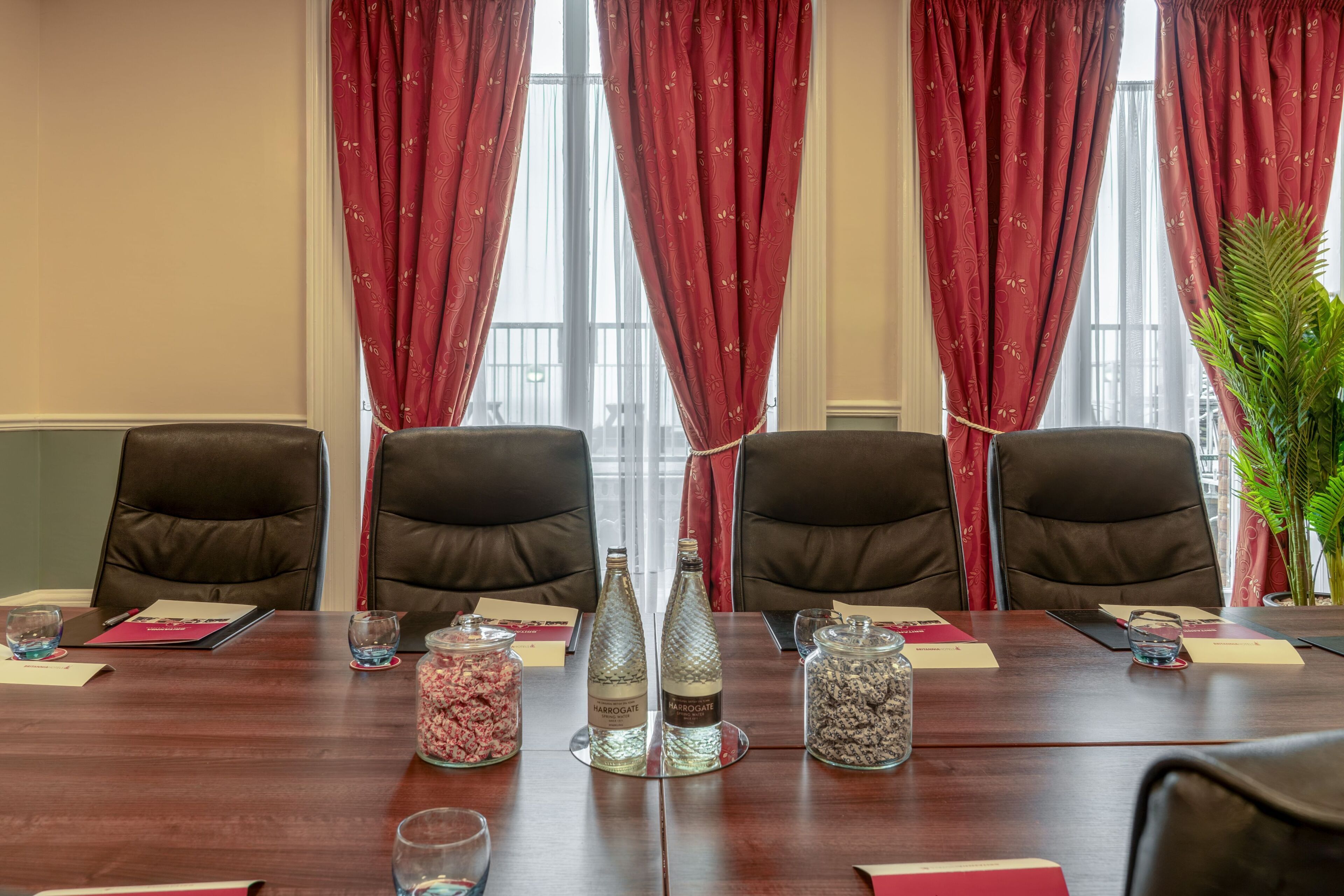 A conference room with a wooden table, four leather chairs, red curtains, and two salt and pepper shakers.