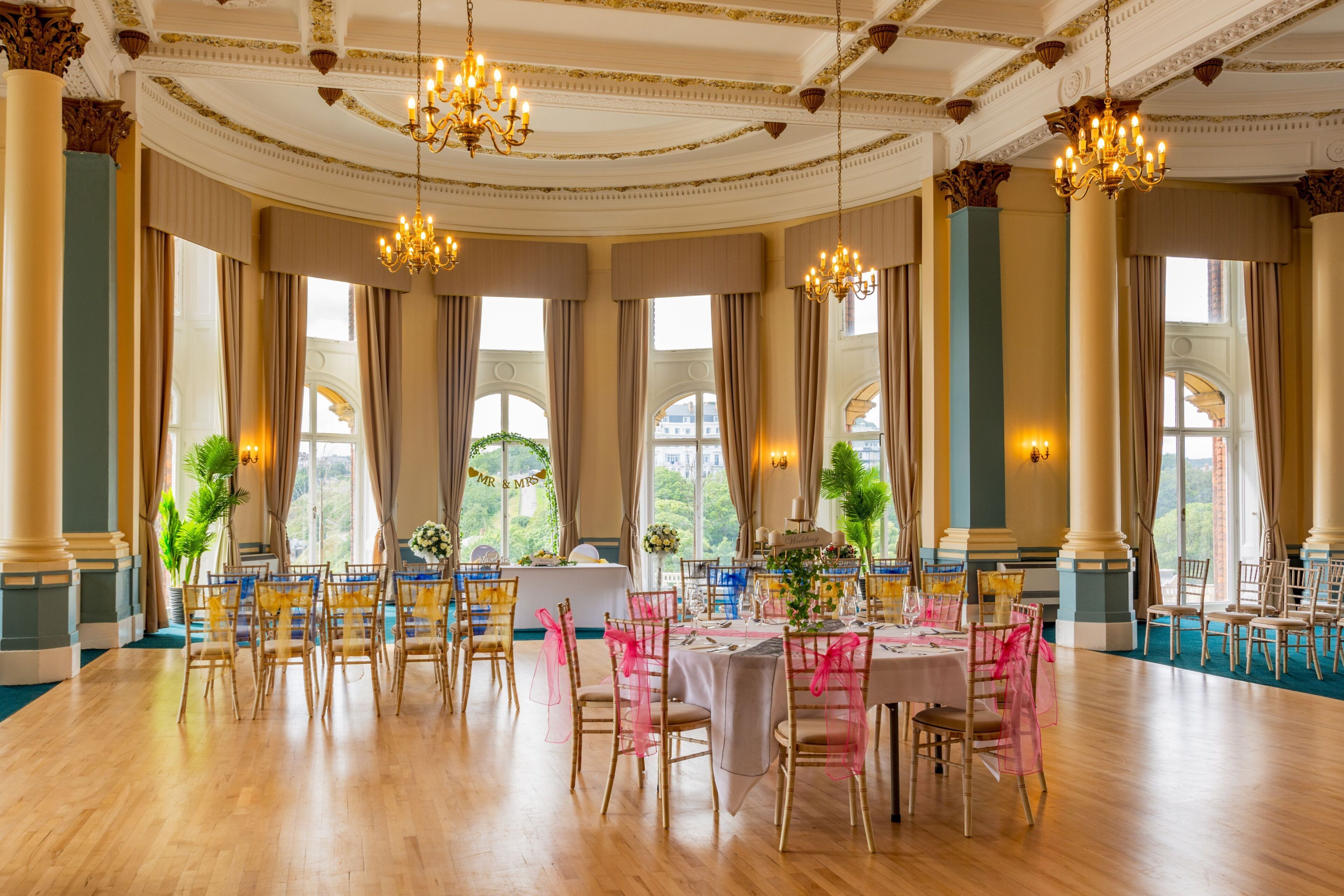 A grand hall with chandeliers, large windows, and a formal dining setup.