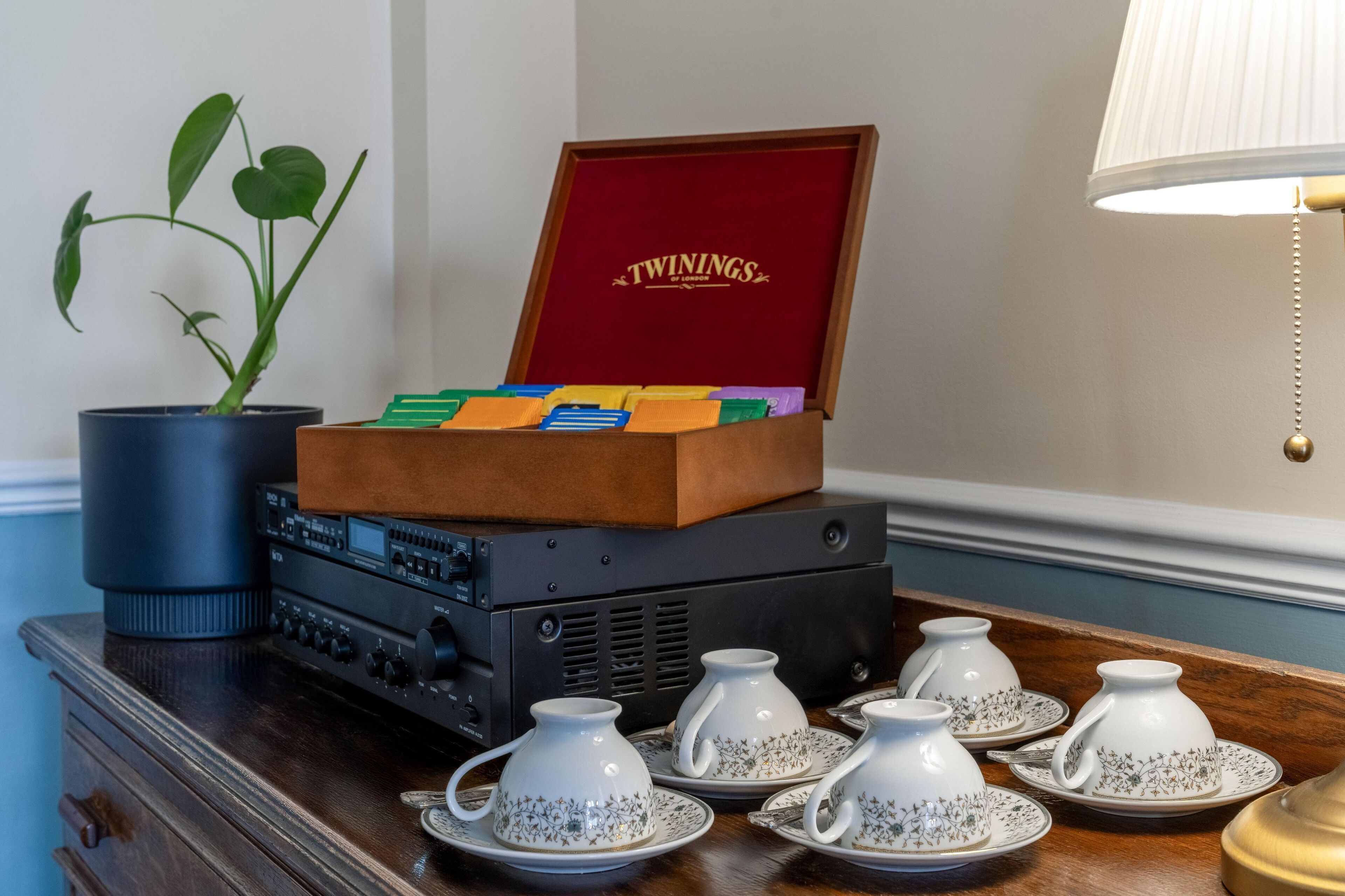 A wooden box with a red interior labeled "TWININGS" containing colorful items, placed on a table with a potted plant and audio equipment.