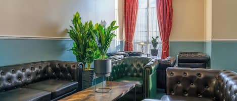 Una sala de estar con un sofá de cuero marrón oscuro, un sillón verde y una mesa de centro de madera. Hay dos plantas en macetas y una lámpara sobre la mesa.