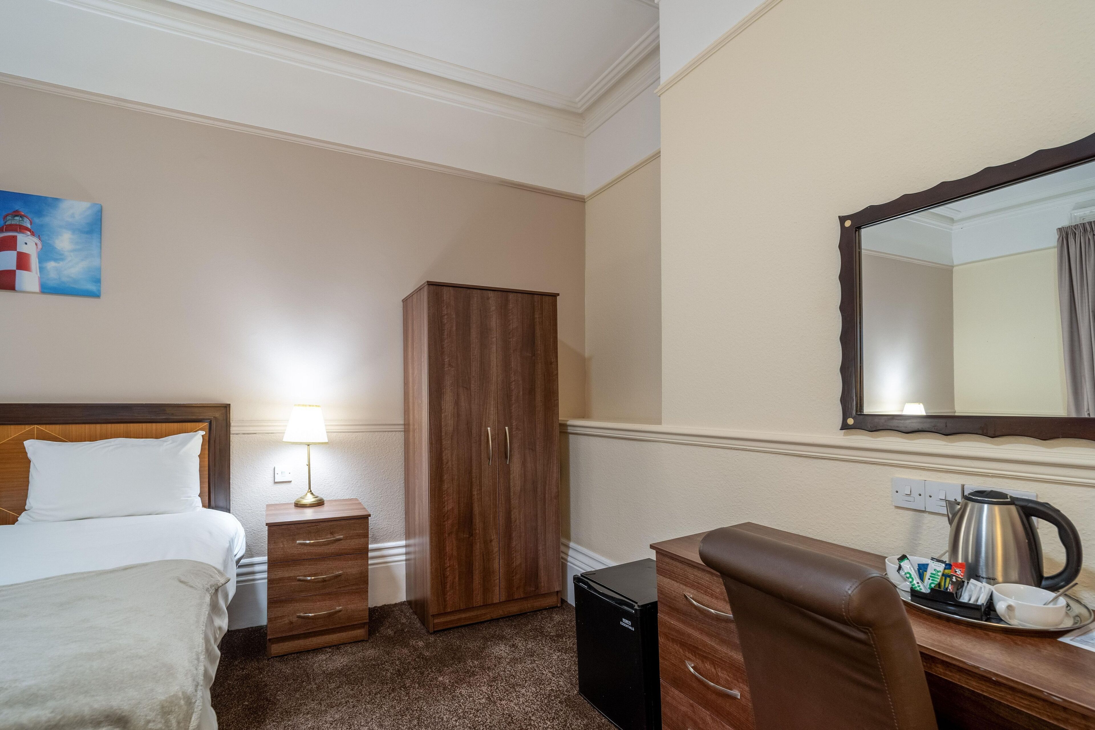 Ein Hotelzimmer mit einem Bett, einem Holzschrank, einem Schreibtisch mit Wasserkocher und Tablett sowie einem Gemälde eines Leuchtturms.