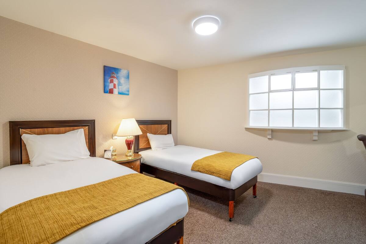 Une chambre d’hôtel avec deux lits, une table de chevet avec une lampe, un tableau au mur et une fenêtre à vitres dépolies.