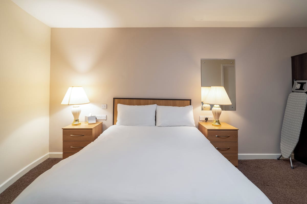 Un lit bien fait, avec du linge de lit blanc, deux tables de chevet avec des lampes, une tête de lit en bois et un miroir fixé au mur.