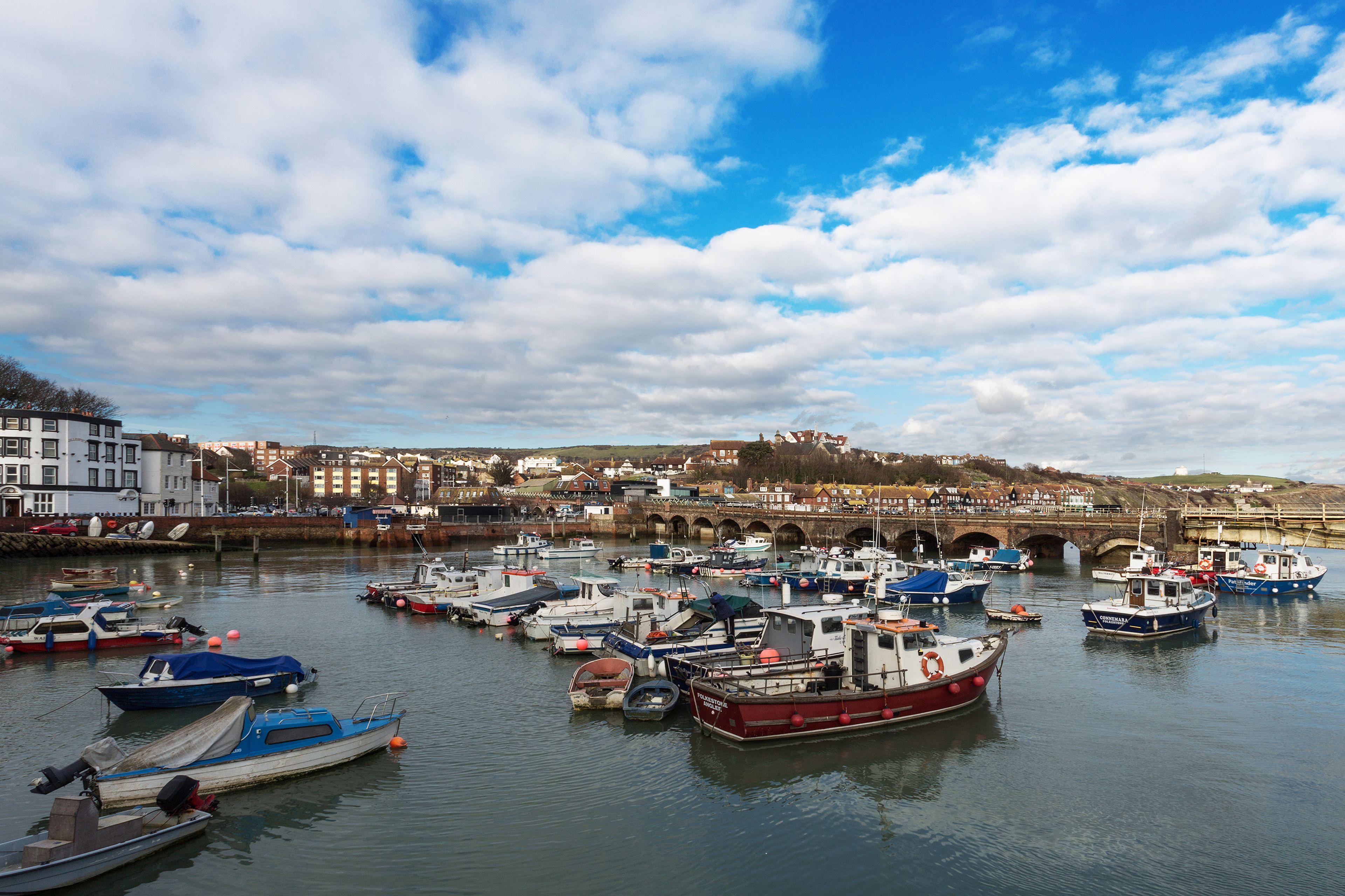 A harbor with multiple boats docked, a bridge in the background, and buildings along the waterfront.