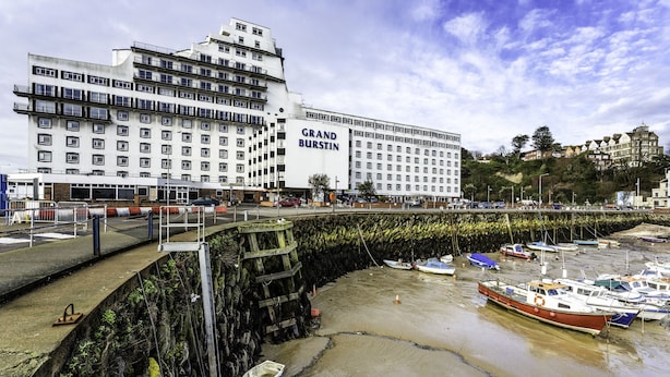 Un hotel gran amb múltiples pisos i balcons, un moll amb embarcacions i vista al mar.