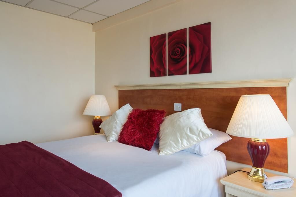 A hotel room with a bed, two bedside lamps, a red throw pillow, and a red rose artwork on the wall.
