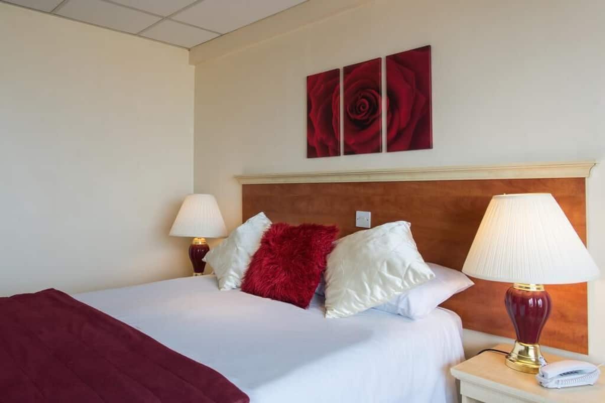 A hotel room with a bed, two bedside lamps, a red throw pillow, and a red rose artwork on the wall.