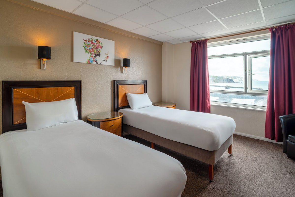 Une chambre d’hôtel avec deux lits, une table de chevet, une lampe, un tableau et une fenêtre avec des rideaux.
