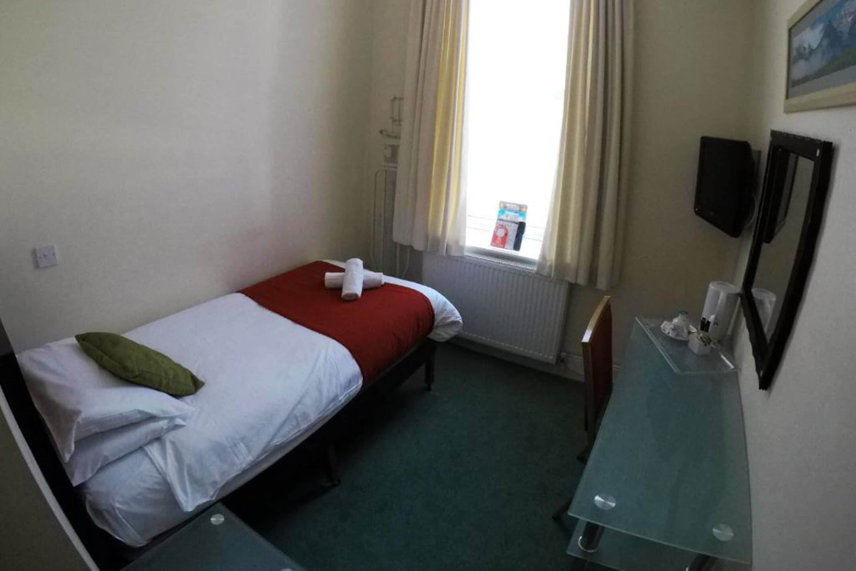 Une petite chambre propre, avec un lit simple, une table de chevet, un miroir et une fenêtre avec des rideaux.