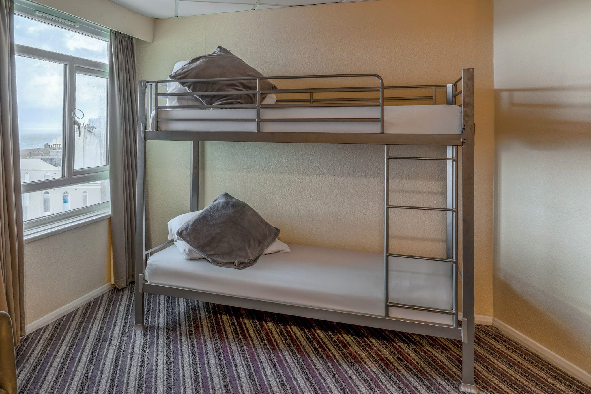 A bunk bed with a window view, a striped carpet, and a pillow on the lower bunk.