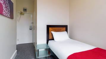 Una habitación de hotel con cama, una mesita auxiliar de vidrio, una colcha roja, un cabecero de madera y una puerta con interruptor de luz.
