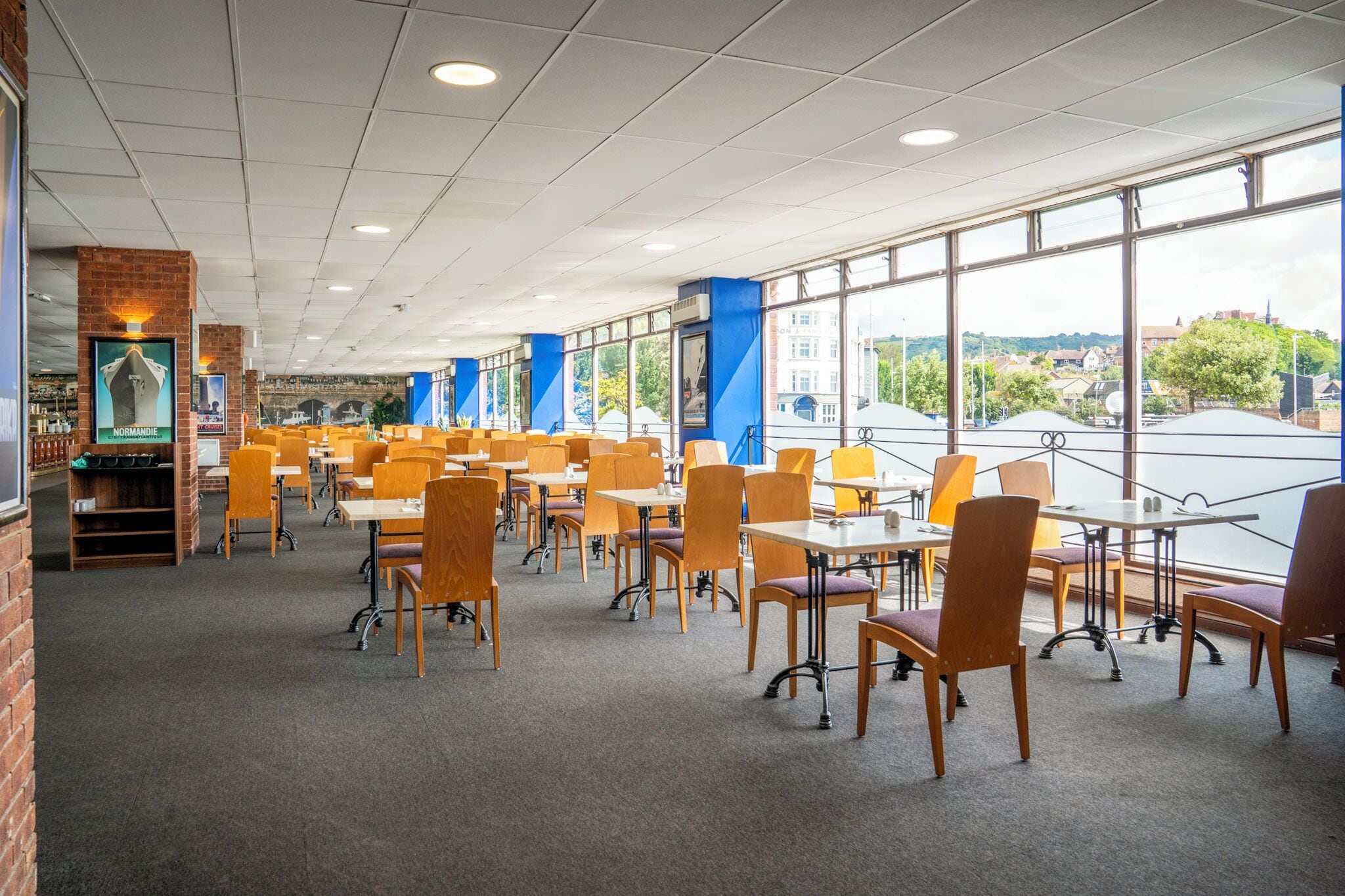 A spacious cafeteria with tables and chairs arranged in rows, large windows offering a view of the outside, and a brick wall on one side.