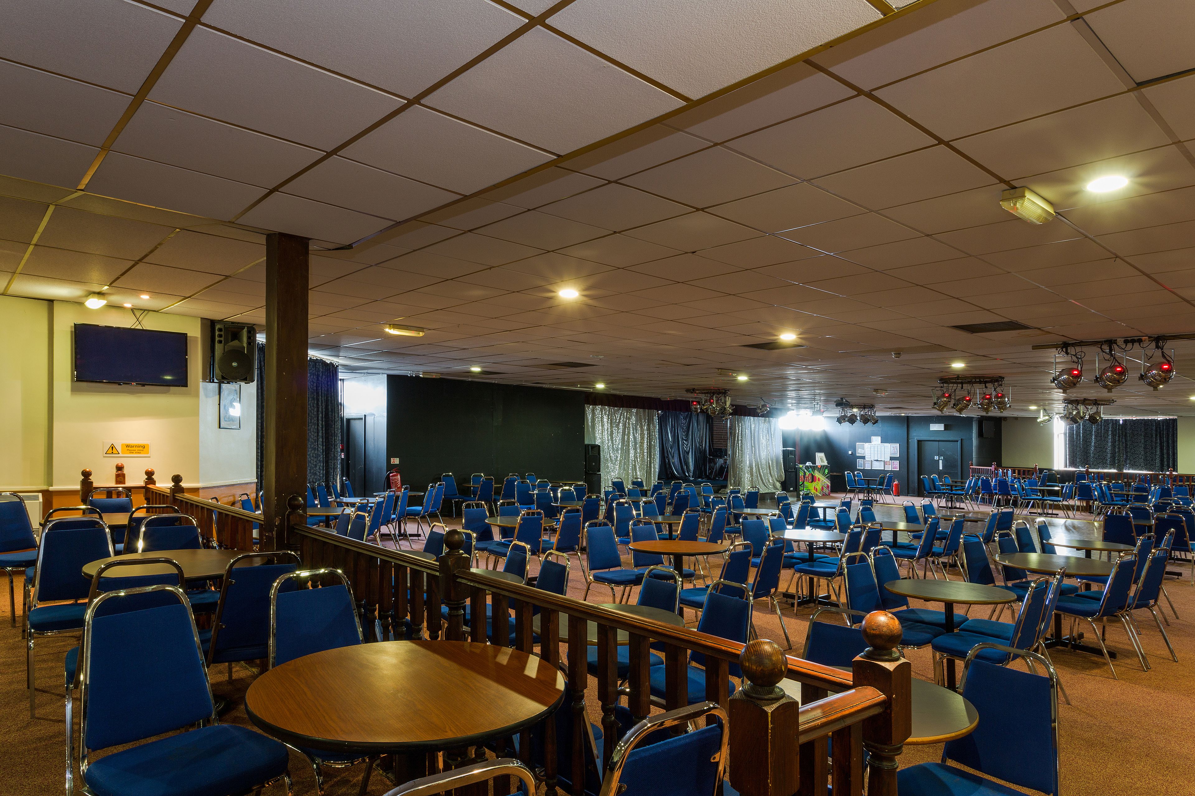 Un salón amplio con filas de sillas y mesas azules, un escenario con cortinas y un sistema de sonido.