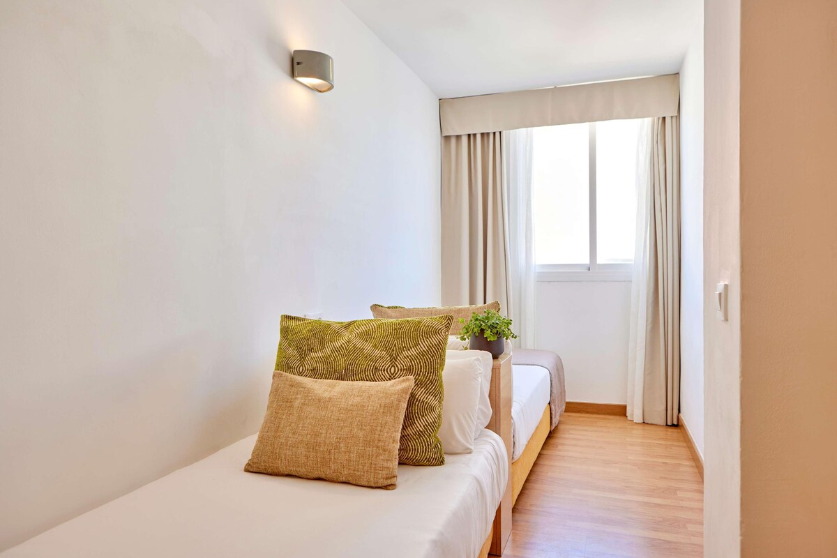 A bedroom with a bed, pillows, a nightstand with a plant, a window with curtains, and a wall-mounted light.