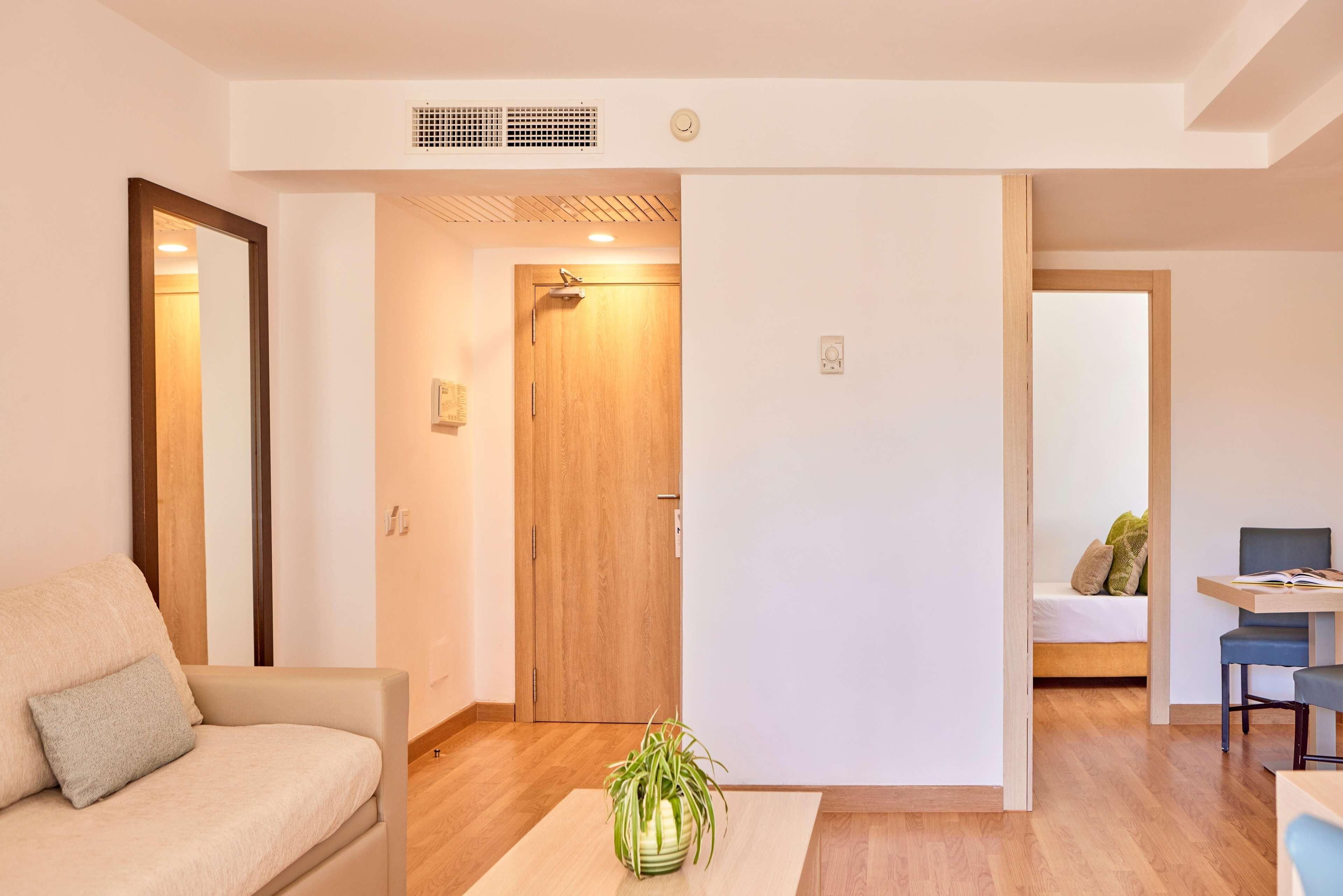 Un salon moderne avec un canapé, une table basse agrémentée d’une plante et une porte en bois donnant sur une chambre.