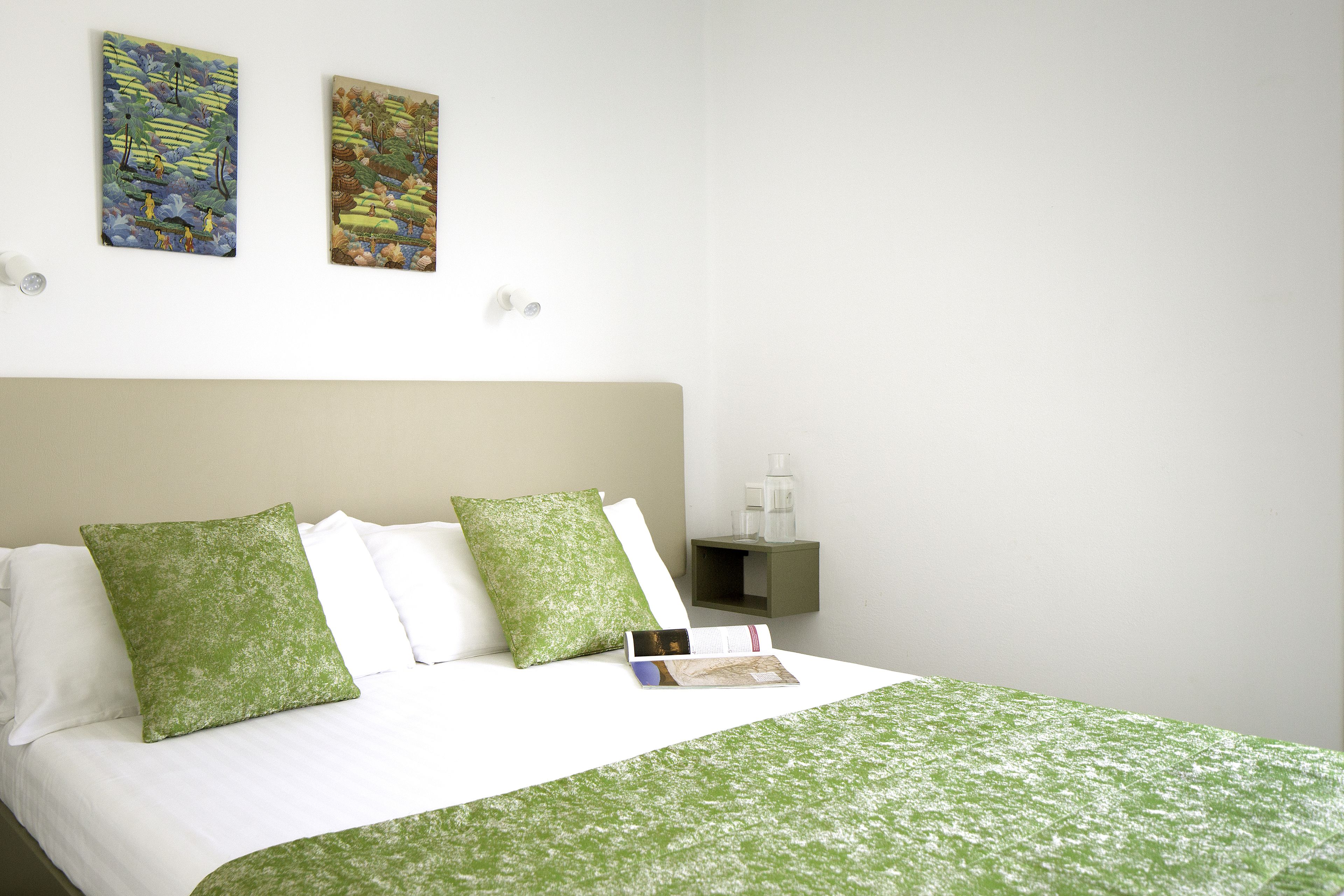 Una cama bien hecha con ropa de cama verde, una mesita de noche con un libro y dos cuadros en la pared.