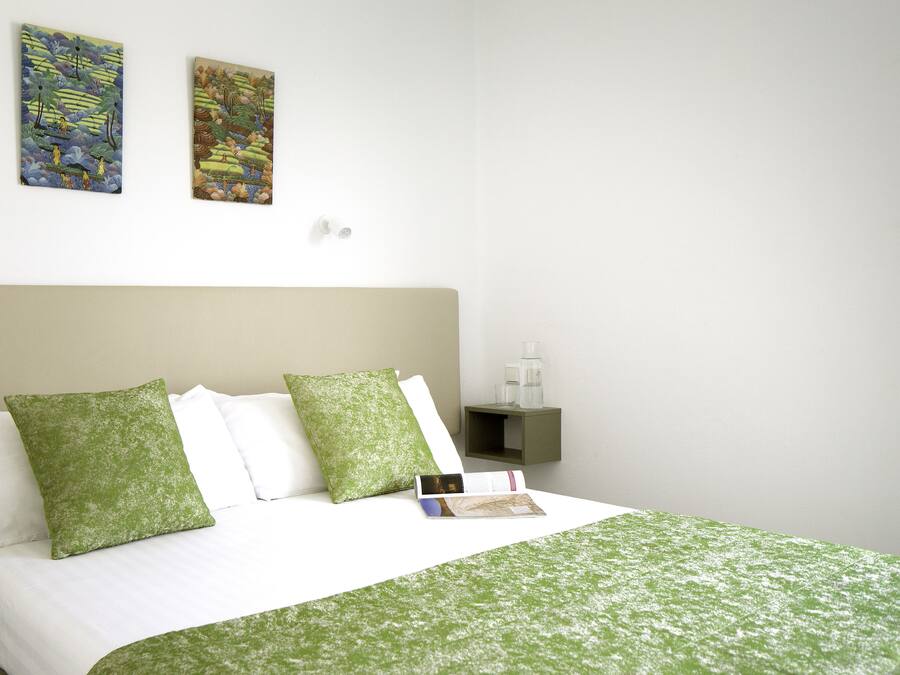 Una cama bien hecha con ropa de cama verde, una mesita de noche con un libro y dos cuadros en la pared.