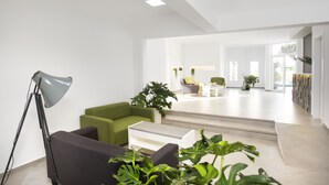 A modern living room with a black sofa, green armchair, and a white coffee table. There is a floor lamp and several potted plants. - El Guarapo Apartamentos (Teguise)