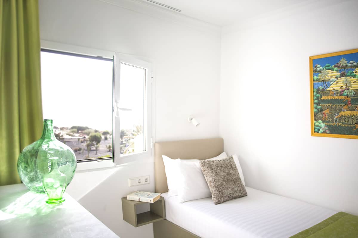A hotel room with a bed, a nightstand, a green vase, and a painting on the wall.