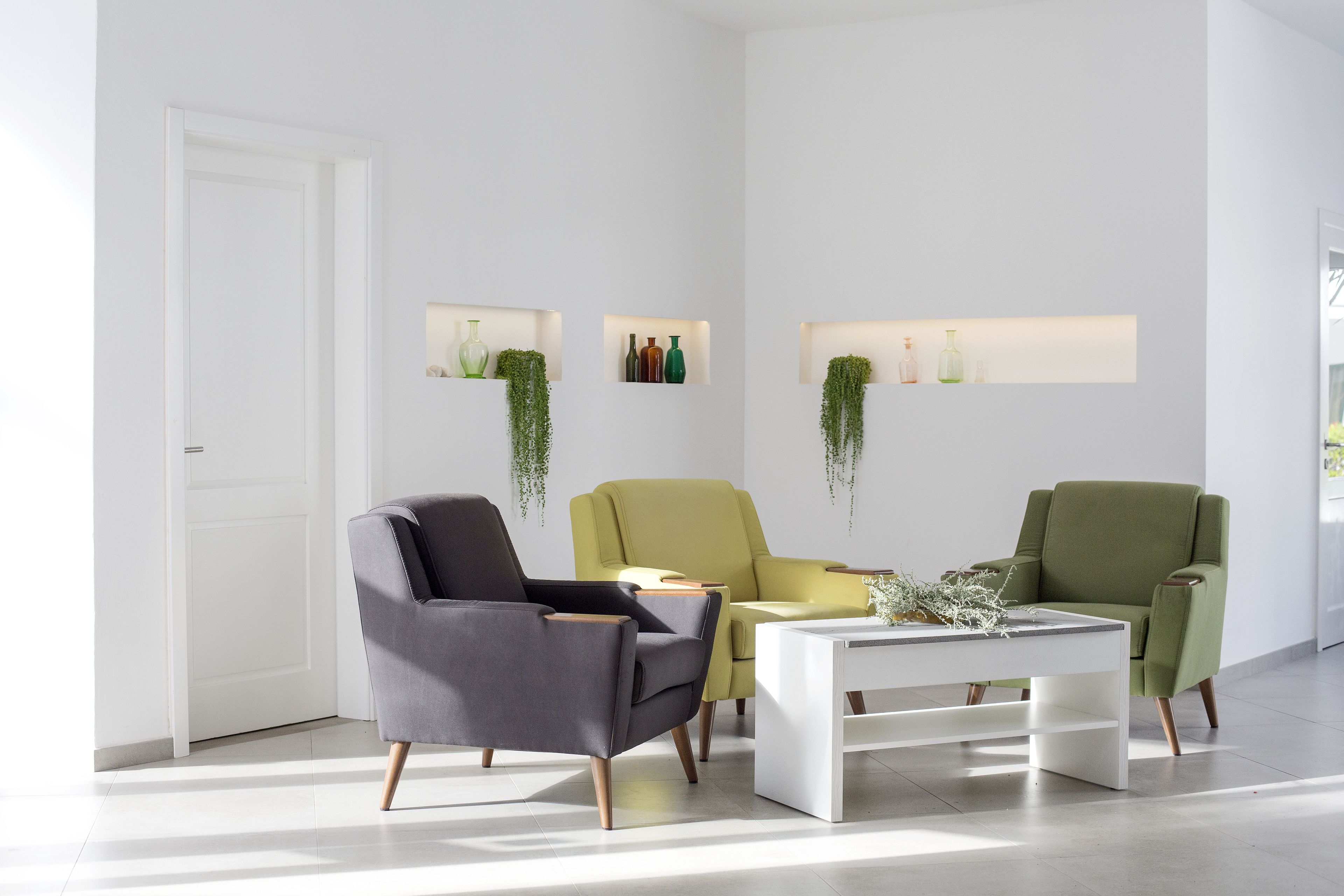 Ein modernes Wohnzimmer mit einer weißen Tür, drei Stühlen, einem weißen Couchtisch und an der Wand befestigten Regalen mit Dekorationen.