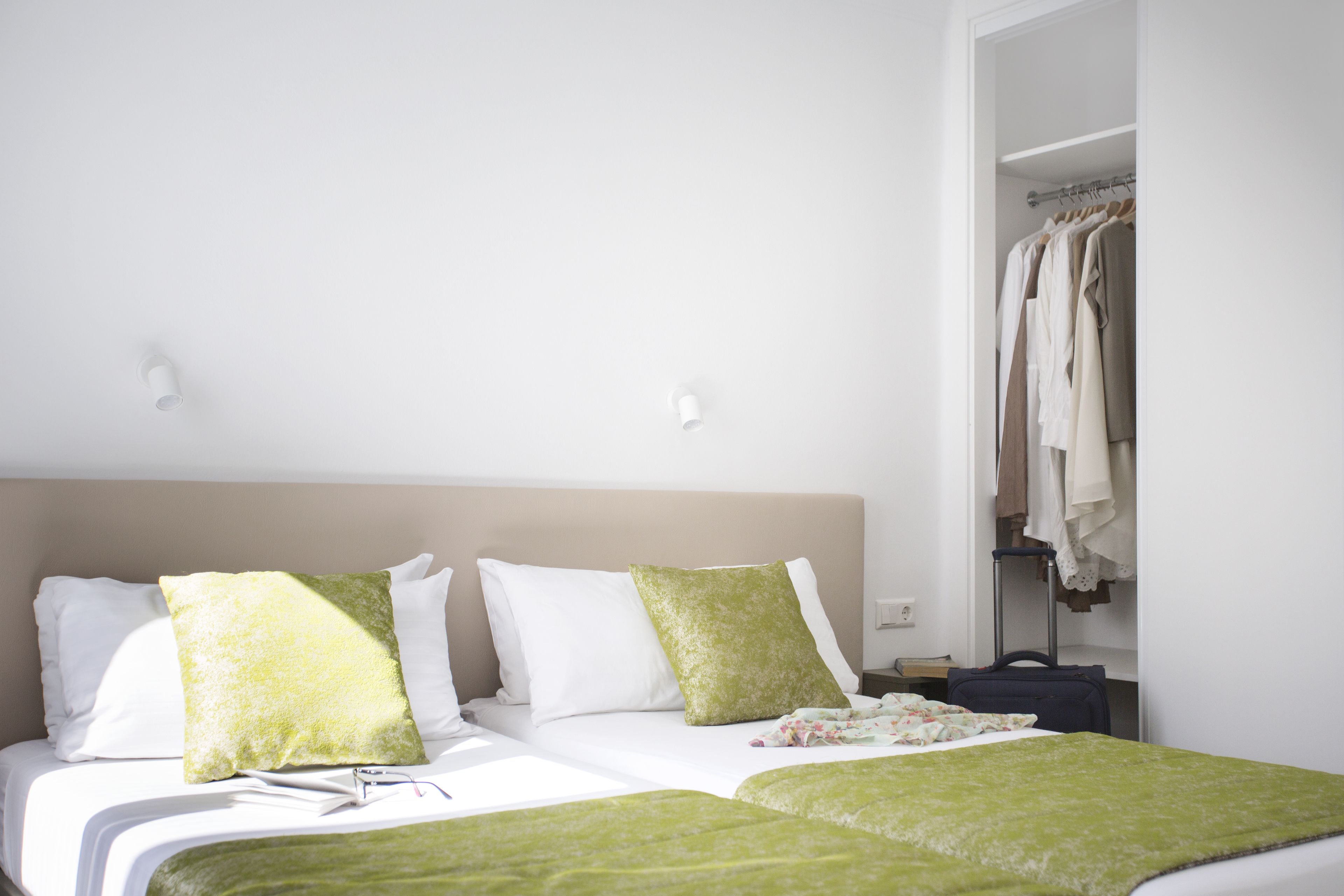 Un lit bien fait, avec une literie blanche et verte, une table de chevet avec une lampe et une armoire où sont suspendus des vêtements.