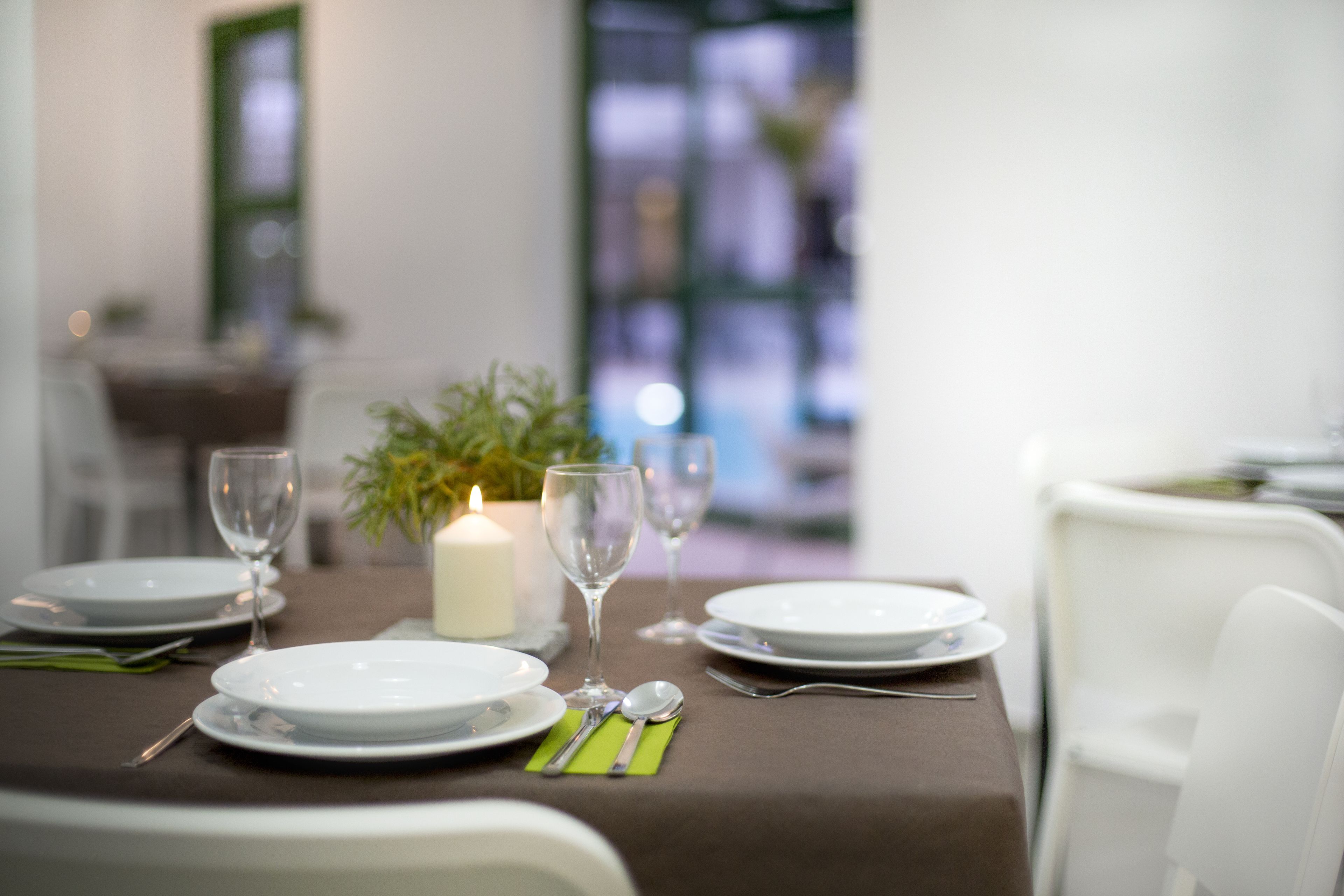 Une table à manger dressée avec des assiettes, des couverts et une bougie, avec une plante et une fenêtre en arrière-plan.
