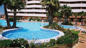 Zona de piscina de hotel con piscina circular, palmeras, sillas de descanso y un edificio de varios pisos al fondo.