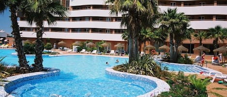 A hotel pool area with a circular pool, palm trees, lounge chairs, and a multi-story building in the background.
