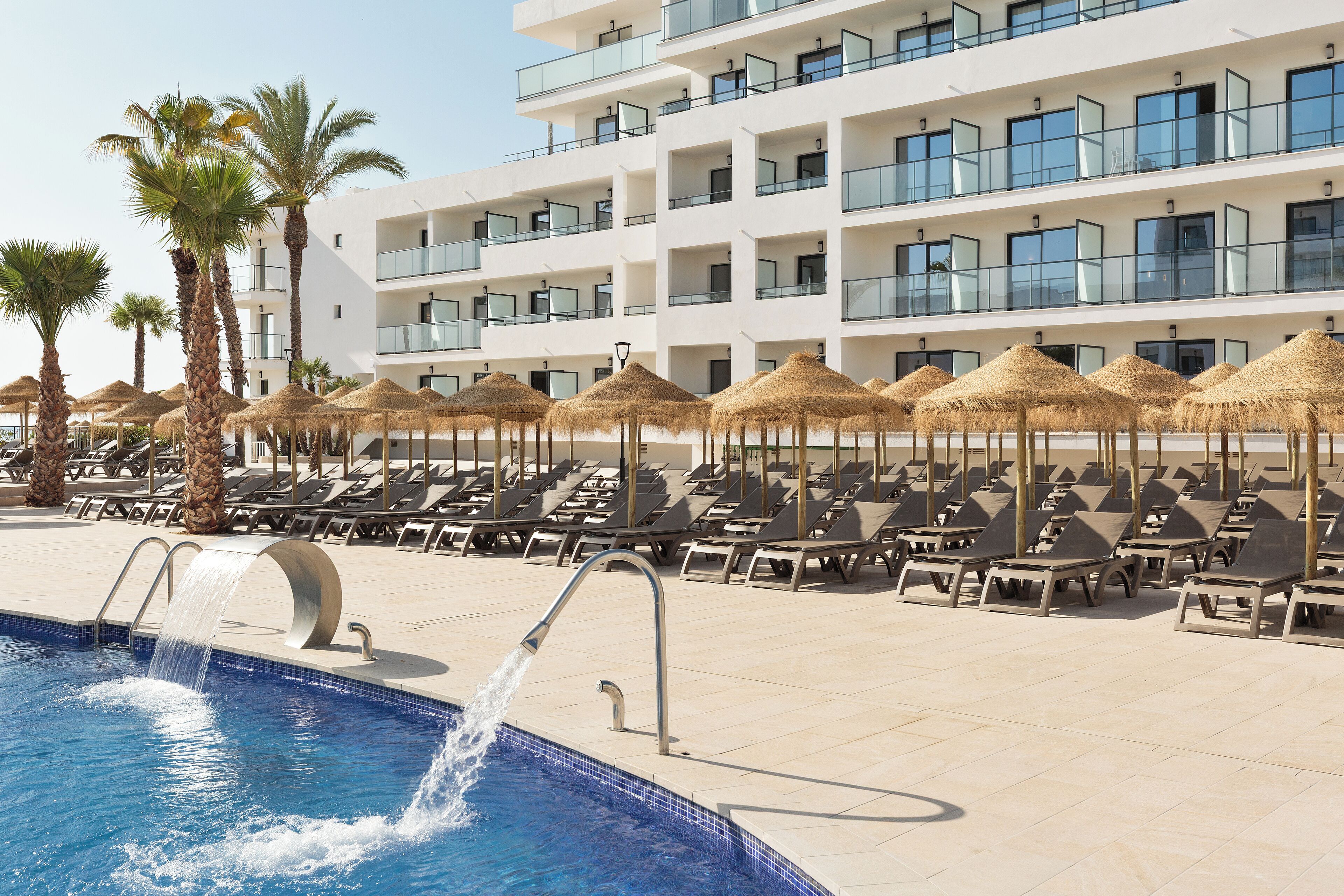 Una piscina con agua que cae de una fuente, rodeada de sillones de descanso y sombrillas de paja, con un edificio de hotel al fondo.