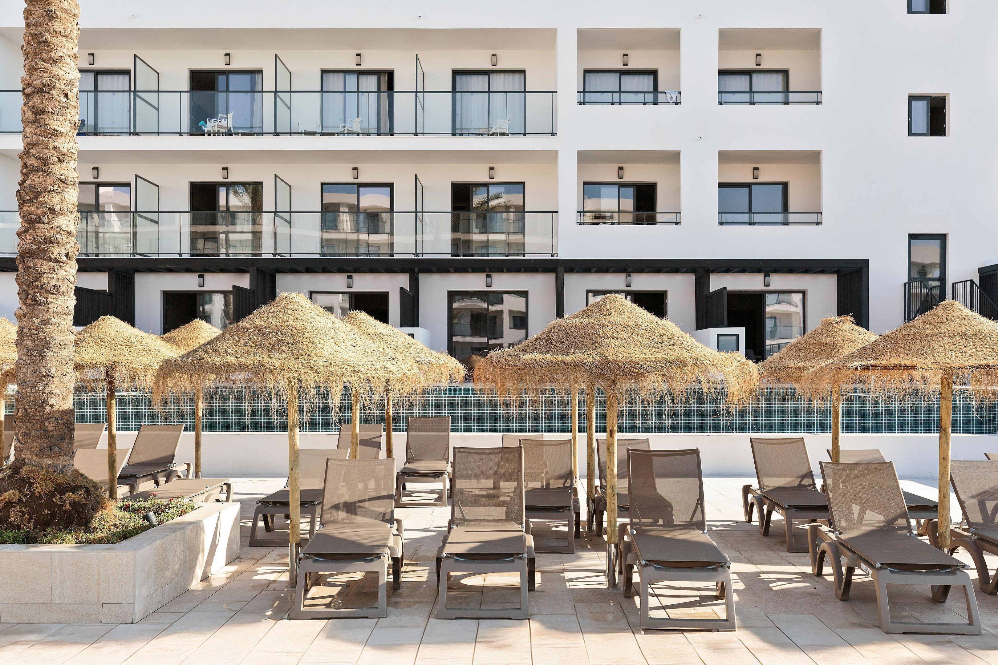 Un hotel con zona de piscina que incluye tumbonas y sombrillas de paja, fachada de edificio moderna y vista despejada a la piscina.