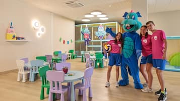 Un área de juegos para niños con sillas y mesas coloridas, un personaje mascota y una estructura de juego con red.