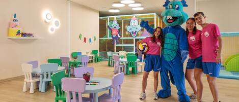 A children's play area with colorful chairs and tables, a mascot, and a netted play structure.