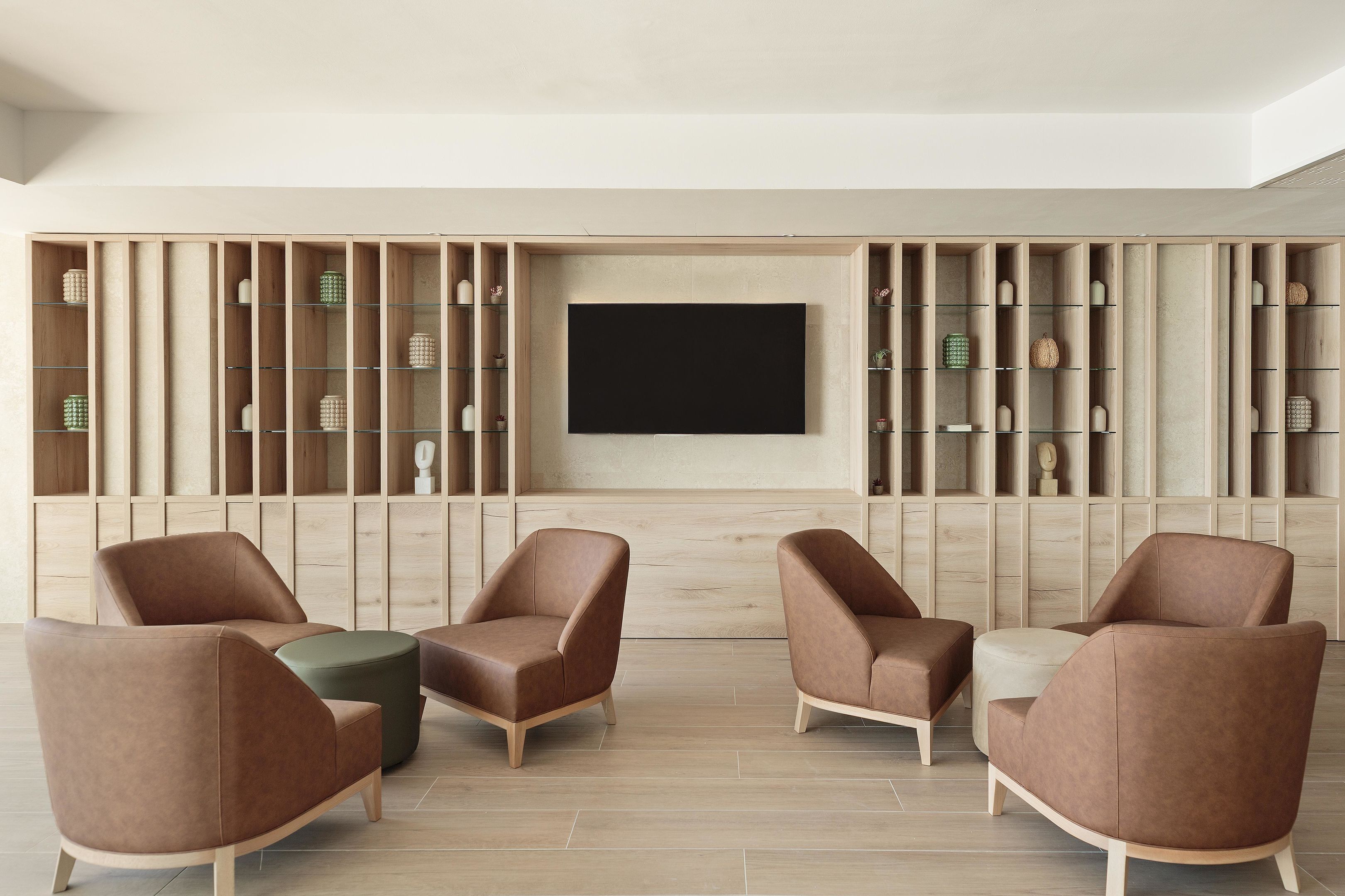 Una sala de estar moderna con un televisor de pantalla plana montado en la pared, rodeada de estanterías de madera con objetos decorativos. Hay tres sillones de cuero marrón y un reposapiés verde.