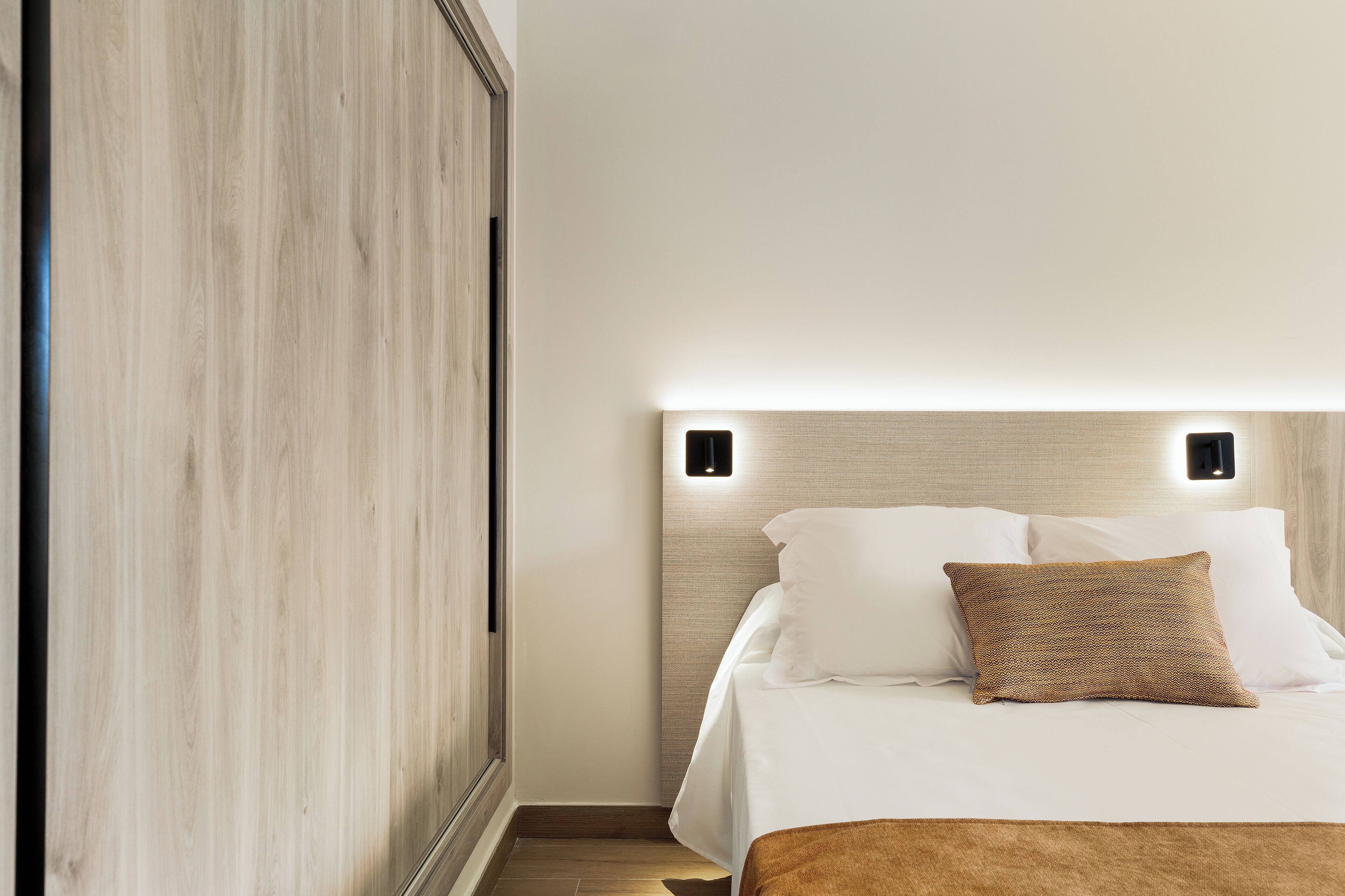 A neatly made bed with white linens and a brown accent pillow, a wooden wardrobe, and a wall-mounted light fixture.